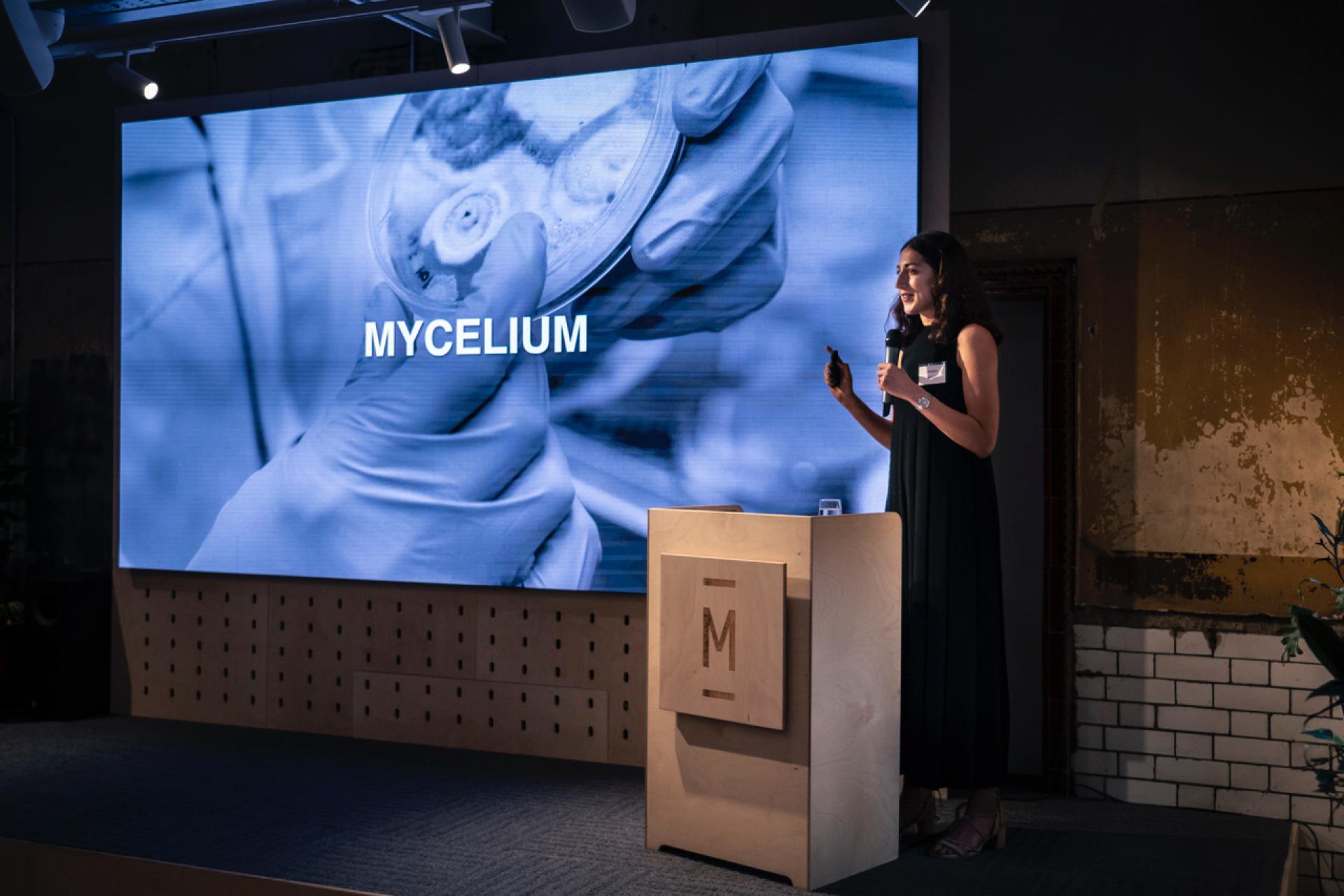 Woman speaking at a presentation with a screen displaying "MYCELIUM," highlighting biomimetic innovations.