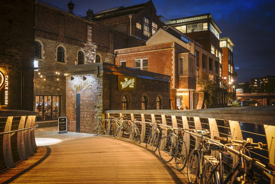 Welcoming entrance of The Wellhead bar in Bristol, featuring cosy lighting, bicycles, and historic architecture at night.