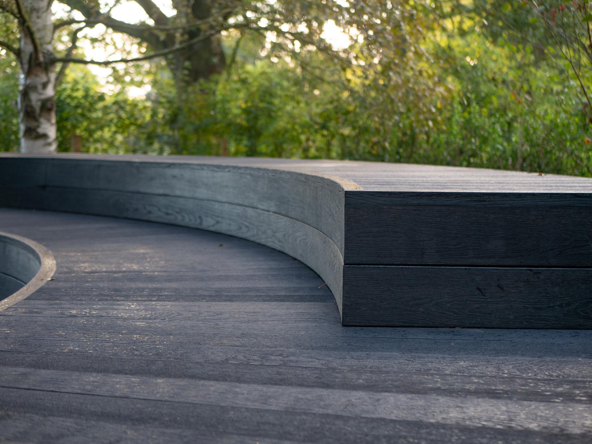 Curved wooden decking made from Millboard's Embered and Burnt Cedar, surrounded by lush greenery at Kew Gardens.