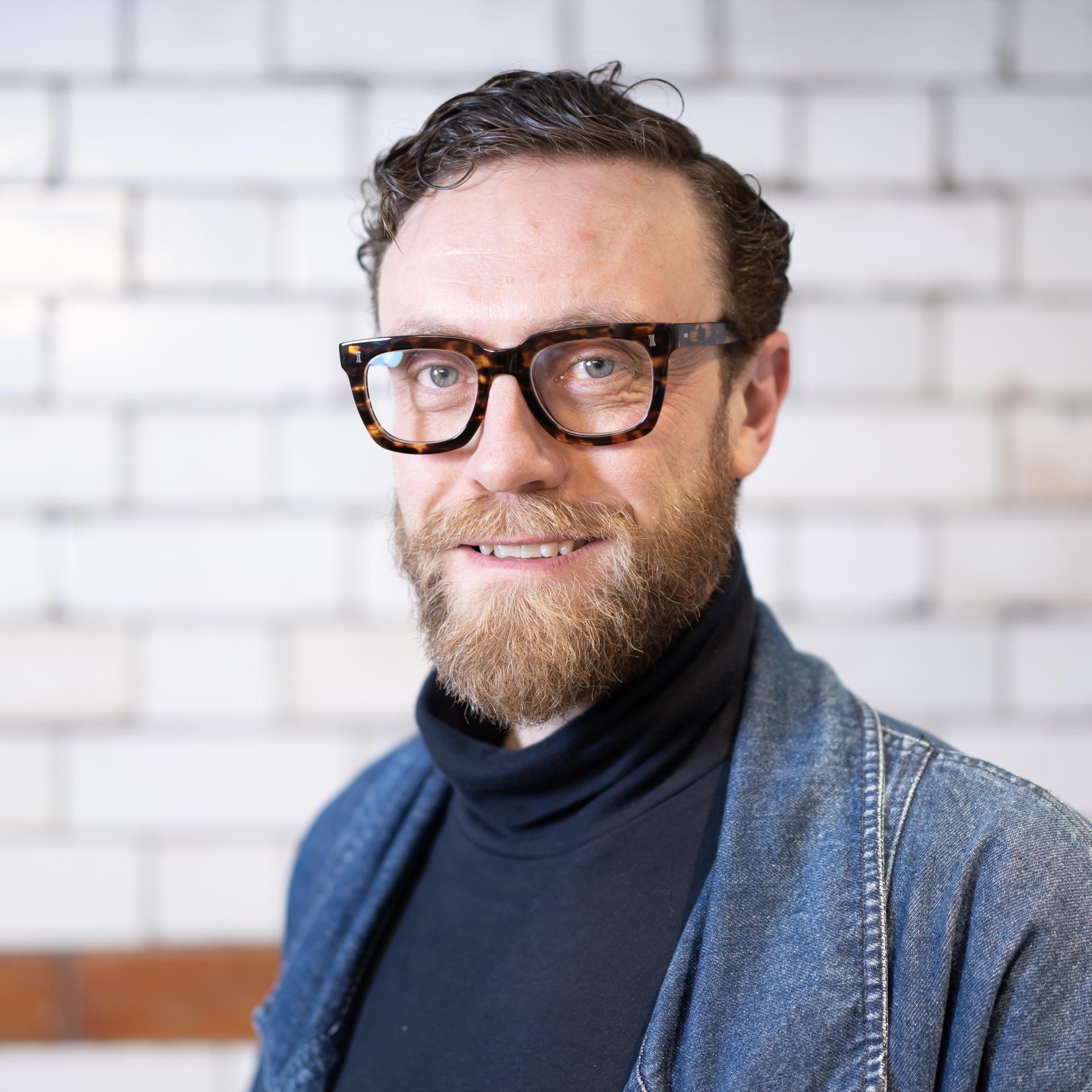 Professional man with glasses and beard, smiling against a modern office background, representing a technologically advanced workplace.