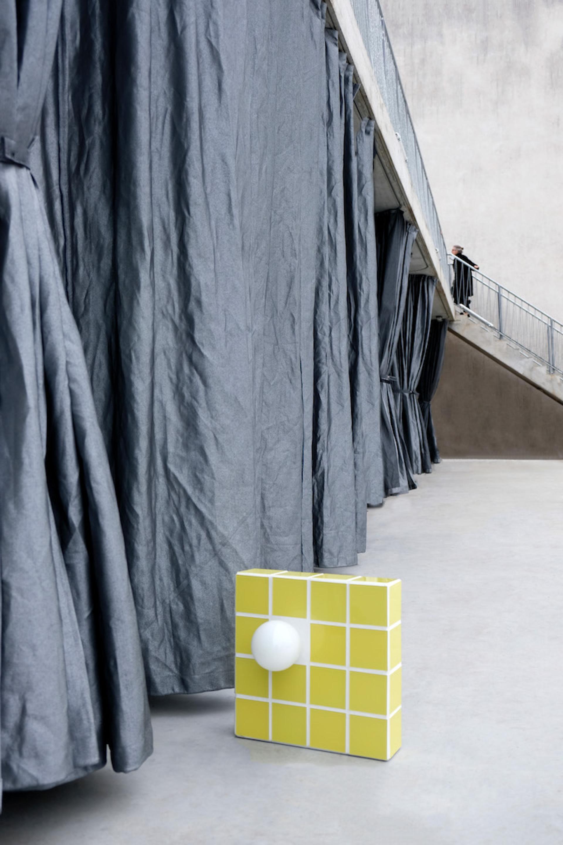 Contemporary German design showcased through a yellow geometric object alongside dramatic gray curtains in an exhibition space.