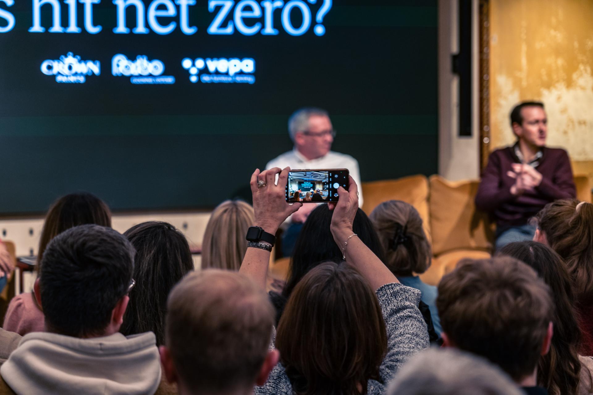 Audience members engage in discussion about achieving Net Zero, with a speaker on stage and people recording the event.