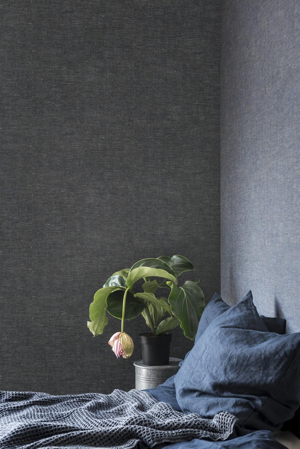 Textured fabric wallcovering with a dark color, complemented by a potted plant and cozy bedding in a stylish interior.