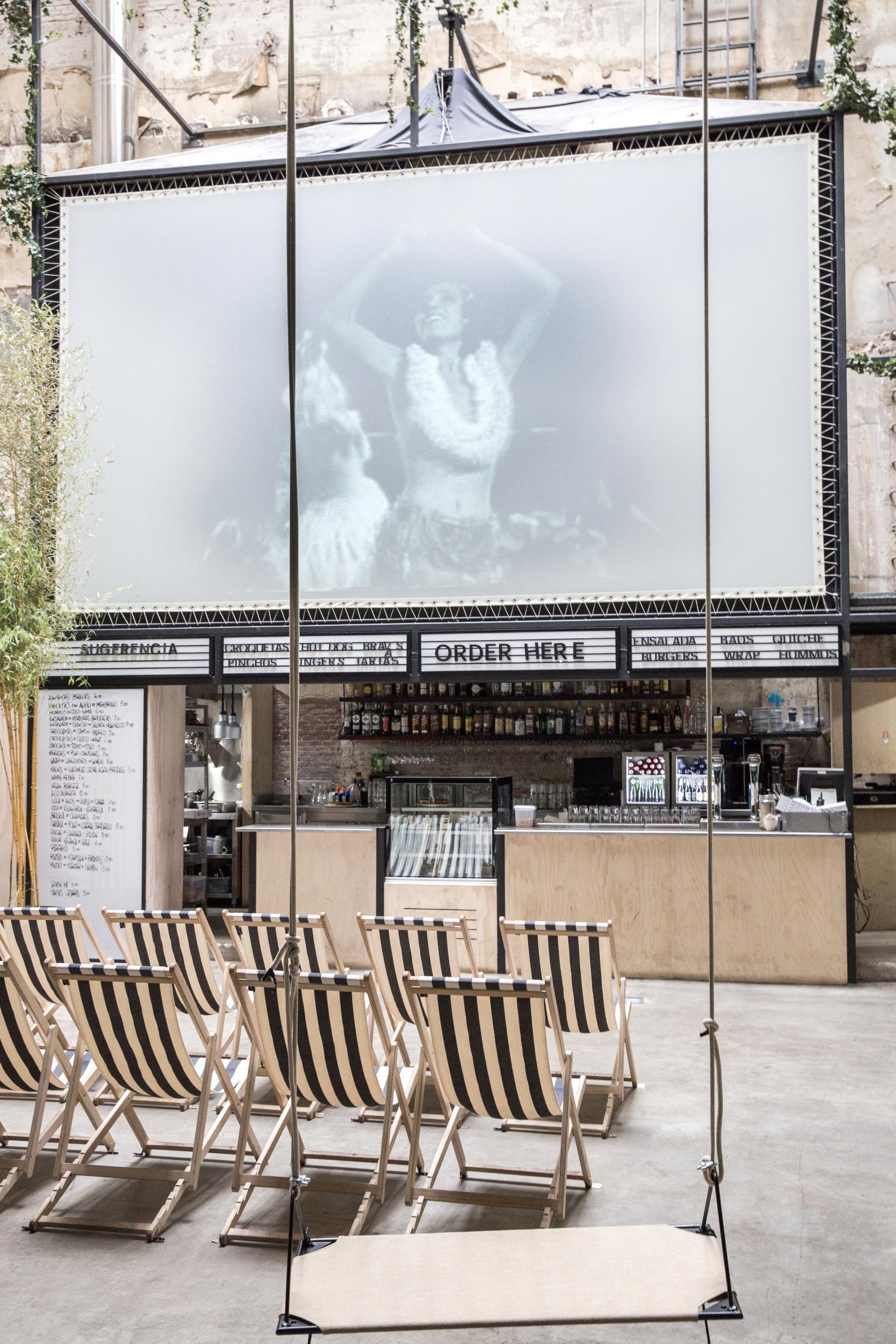Indoor cinema featuring striped deck chairs, a large screen displaying a black-and-white film, and a stylish bar area.