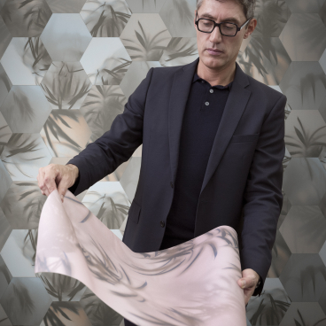 Man in a suit holding a roll of decorative wallcovering, showcasing Tektura Wallcoverings’ new collection against a hexagonal-patterned background.