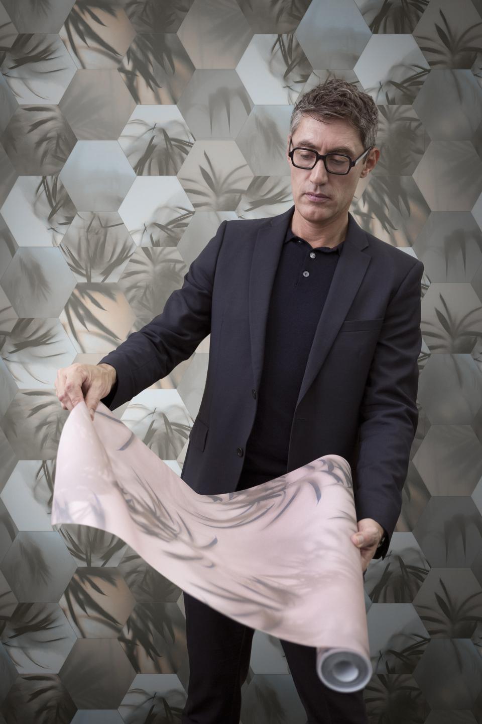 Man in a suit holding a roll of decorative wallcovering, showcasing Tektura Wallcoverings’ new collection against a hexagonal-patterned background.