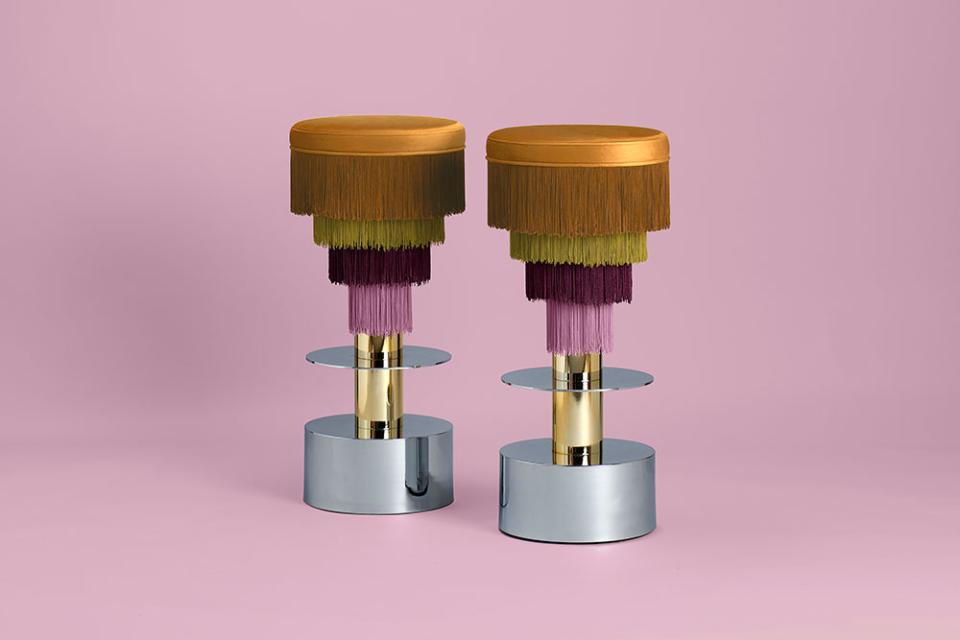 Colorful decorative stools with fringed upholstery, designed by Masquespacio, showcased against a soft pink background.