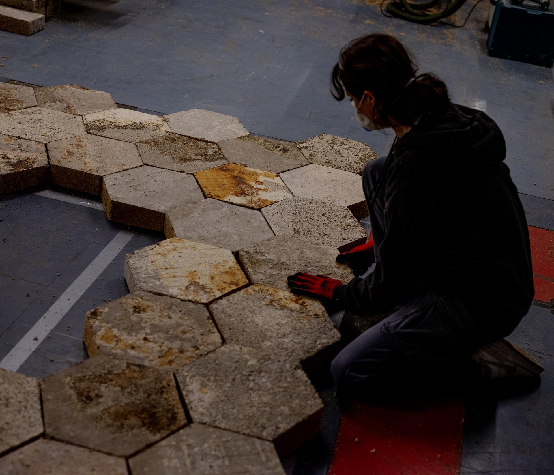A person assembling hexagonal concrete blocks on the floor, contributing to the innovative design for Glastonbury Festival's Hayes Pavilion.