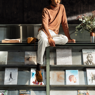 Young man in a brown turtleneck sits on a shelf surrounded by design magazines in a co-working space.
