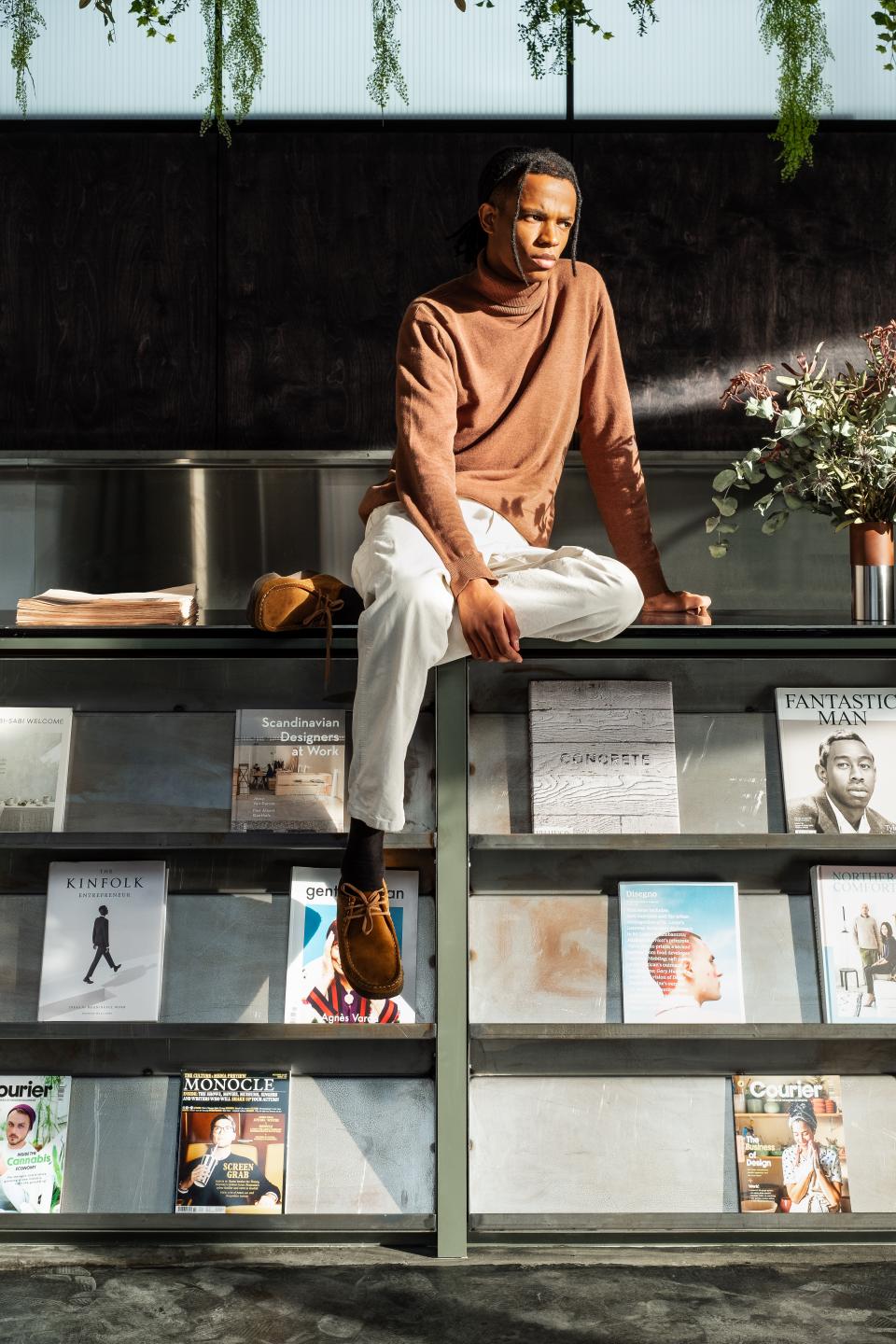 Young man in a brown turtleneck sits on a shelf surrounded by design magazines in a co-working space.