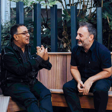 Jon Humphreys and Atul Bansal engaging in conversation on a wooden bench surrounded by greenery, reflecting community and creativity.