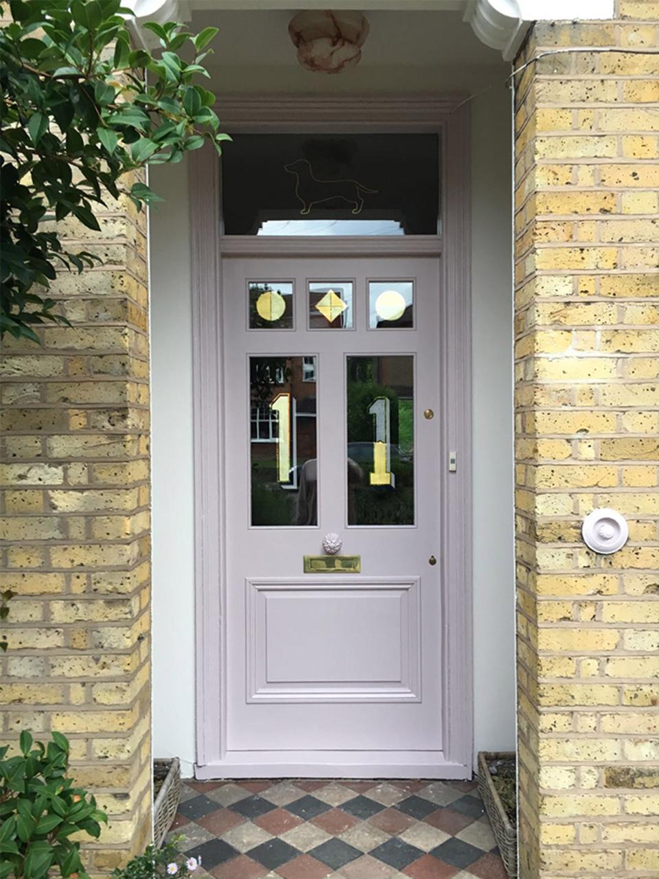 Distinctive pastel pink front door with geometric window designs, framed by exposed brick and greenery in a residential setting.