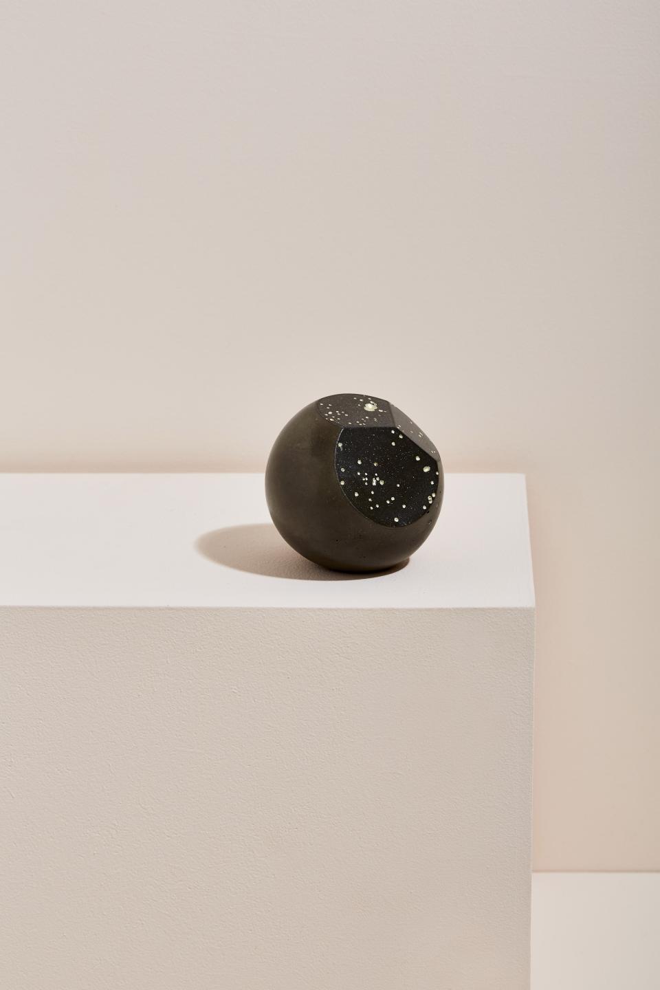 A round abstract sculpture with a speckled black surface, resting on a minimalist white pedestal.