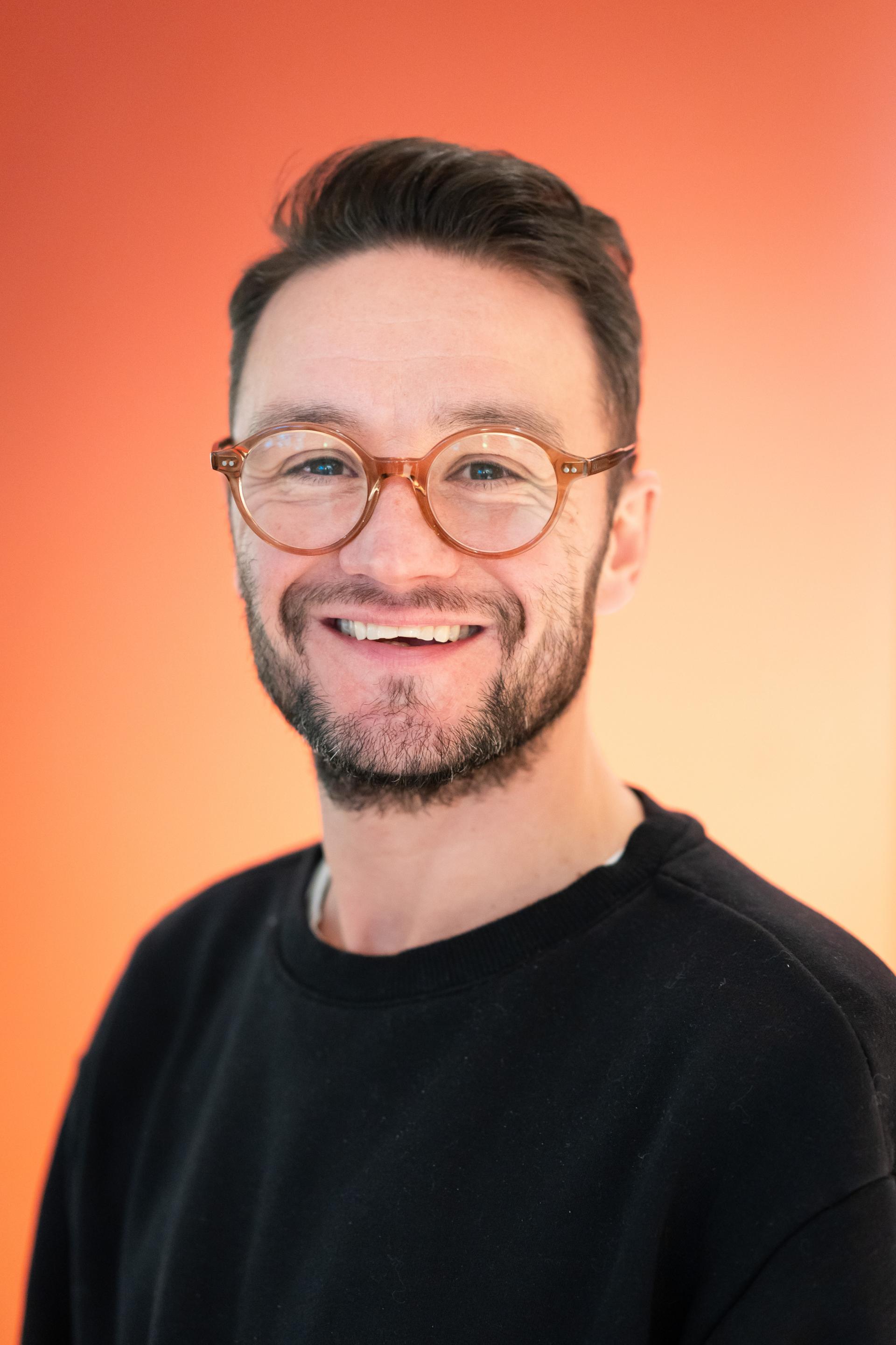 Smiling man wearing glasses in front of a vibrant orange background, embodying workplace flexibility and positivity.