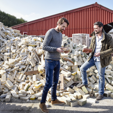 Co-founders of StoneCycling discuss sustainable brick production amid a large pile of recycled materials.