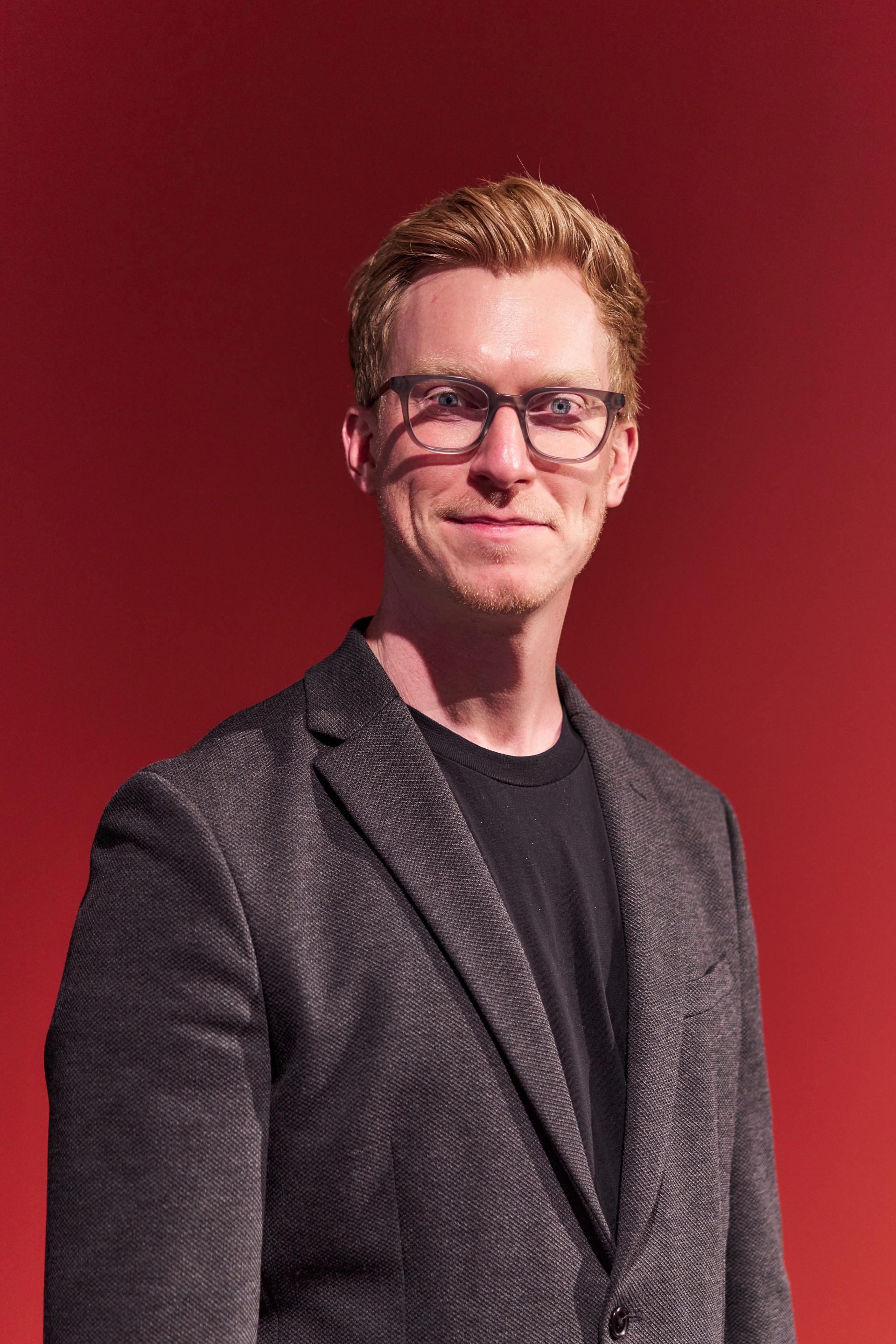 A man in a black shirt and gray blazer poses confidently against a red backdrop at a Glasgow event.
