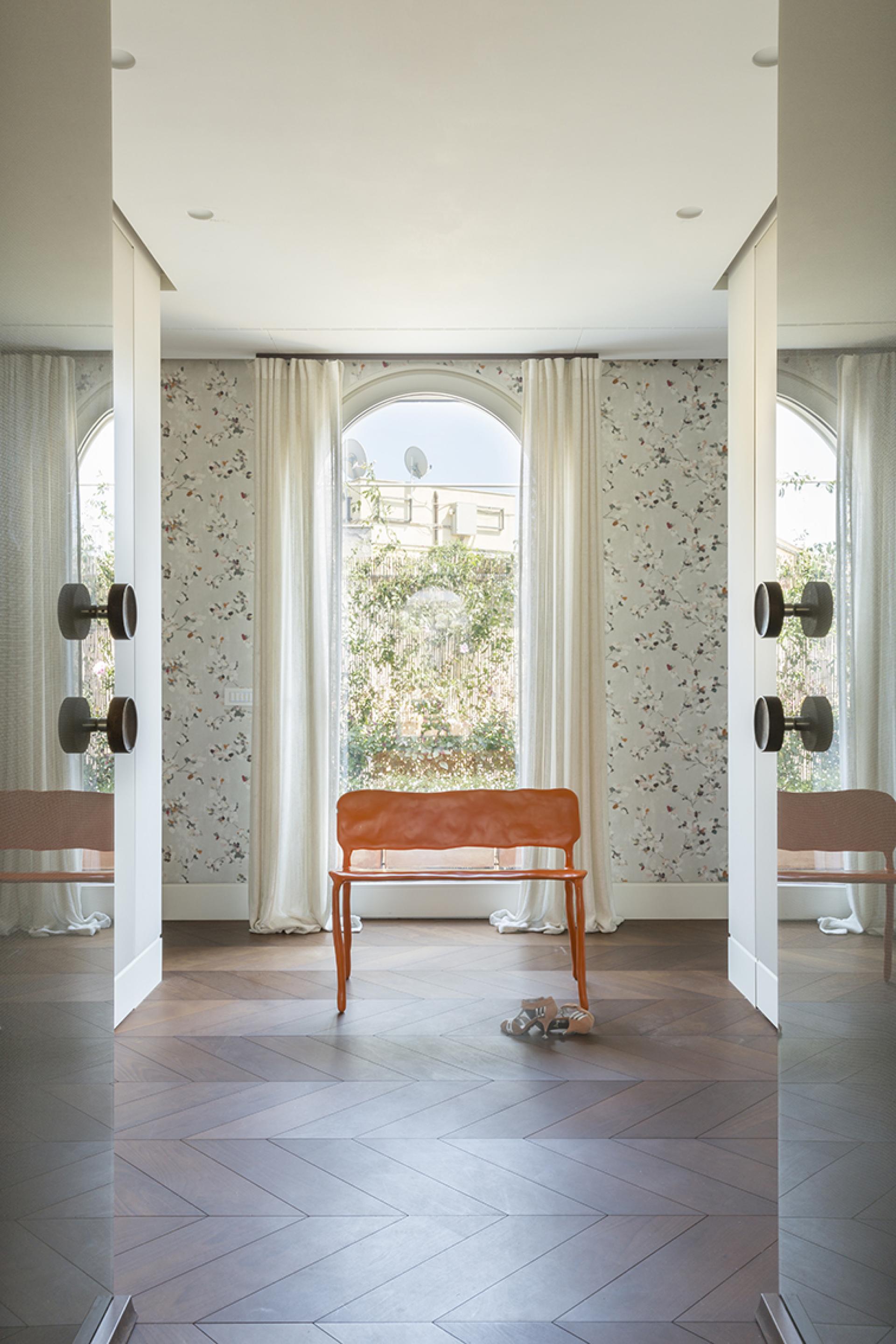 Bright artist's loft interior featuring an orange chair, floral wallpaper, and large windows with garden views in Rome.