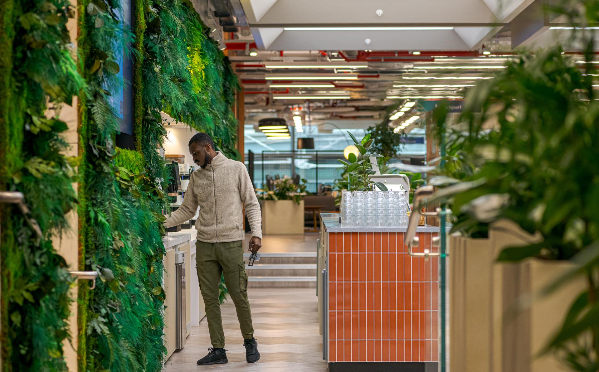 Modern office kitchen with vibrant greenery, showcasing a man preparing coffee in a collaborative workspace.