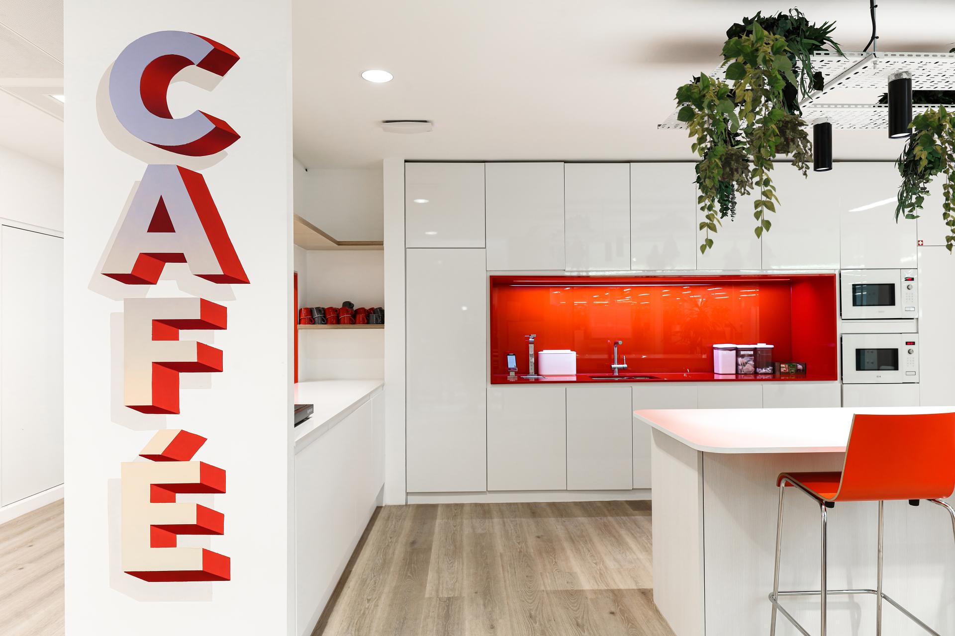 Bright café area in modern office design, featuring red accents, sleek cabinetry, and hanging greenery for low embodied carbon workspace.