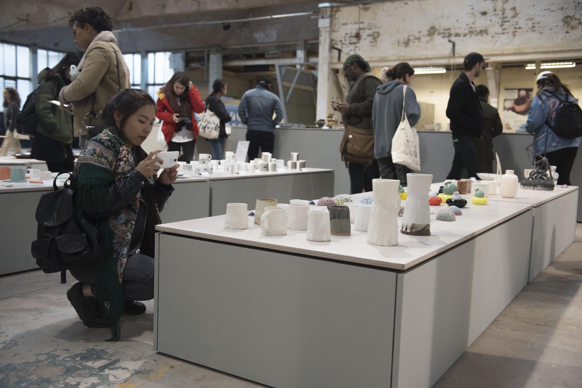 Visitors explore Tana West's (UN)WOVEN installation, engaging with ceramic artworks displayed on modern tables.