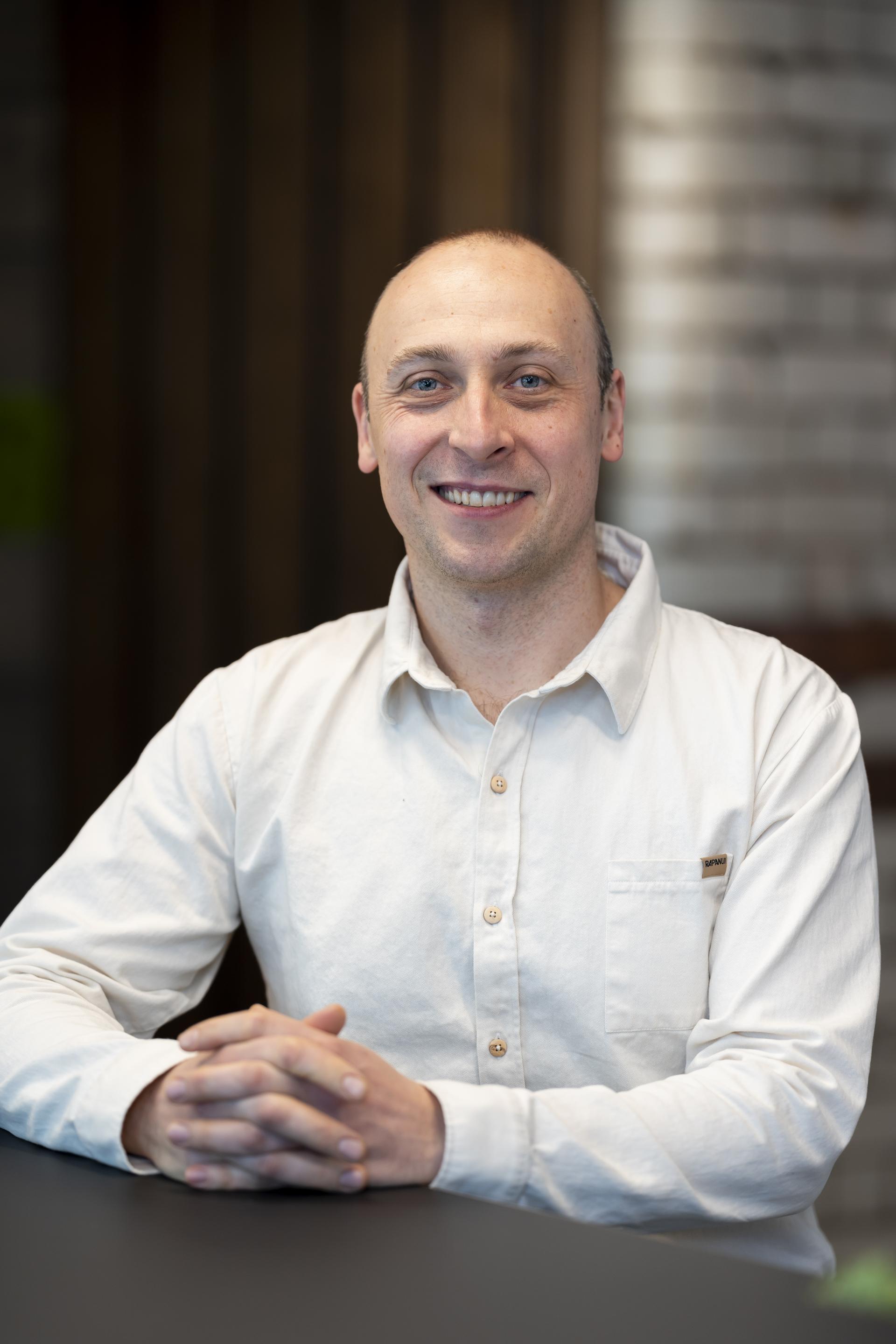 Smiling man in a white shirt sits at a table, discussing technology's role in sustainable design.