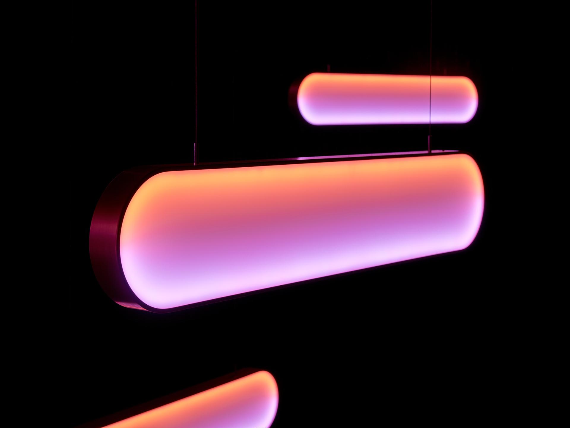 Sleek, elongated pendant lights emit vibrant pink and purple hues, highlighting contemporary design trends at the 2023 DDW.