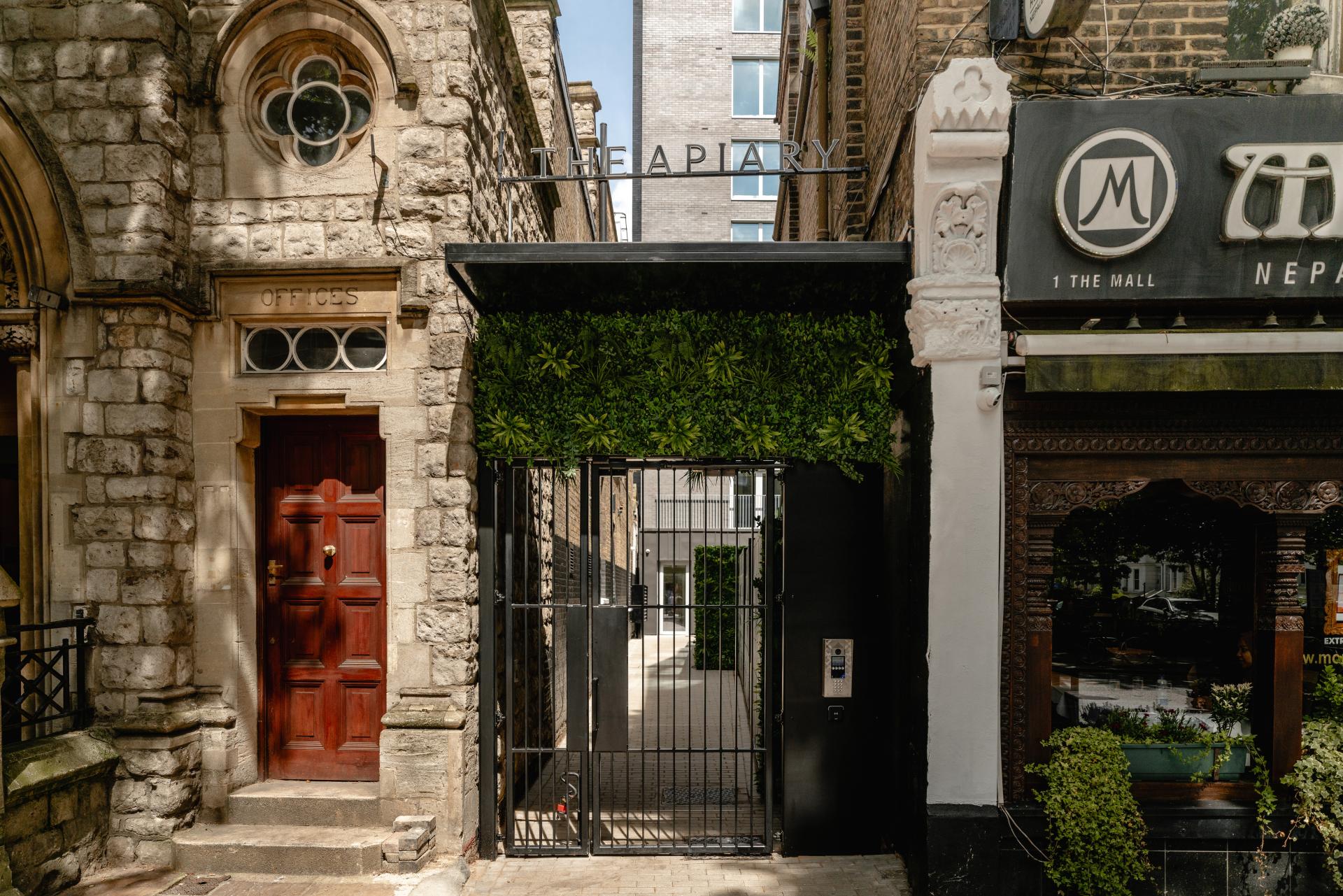 The Apiary entrance featuring a modern gate and greenery, surrounded by historic architecture in London.