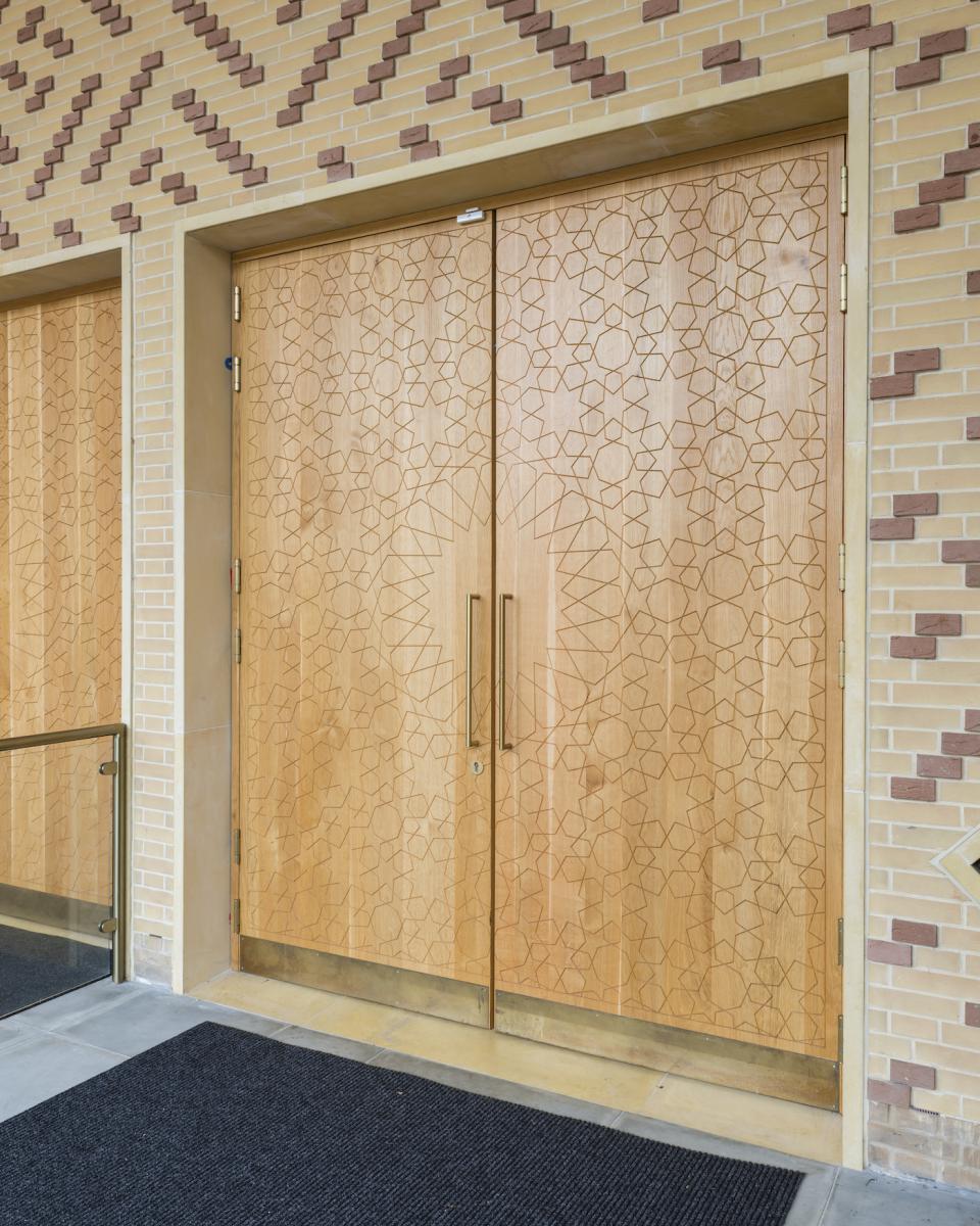 Intricately carved wooden doors of Cambridge Central Mosque, showcasing traditional Islamic patterns against a contemporary architectural backdrop.
