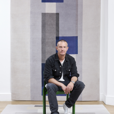 Jonathan Saunders seated on a green chair, showcasing his new rug collection featuring bold geometric patterns and colors.