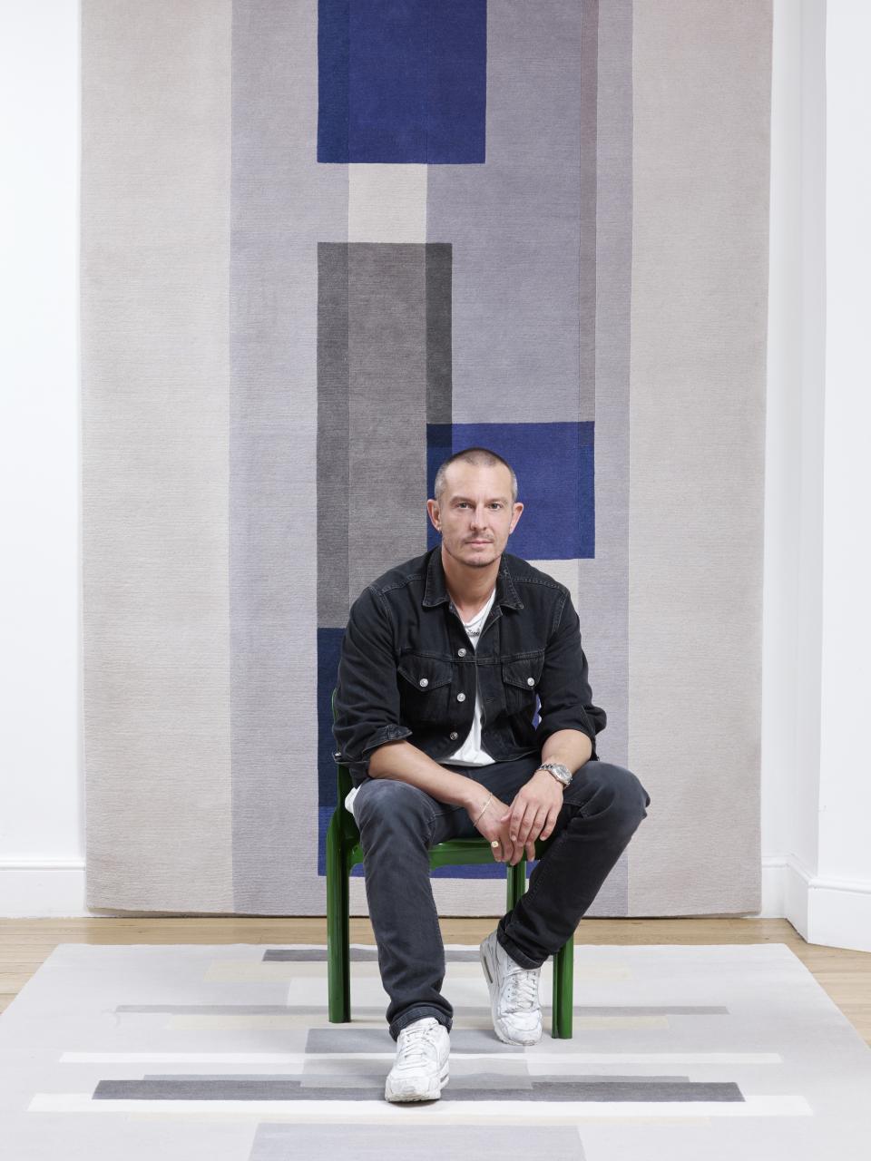 Jonathan Saunders seated on a green chair, showcasing his new rug collection featuring bold geometric patterns and colors.