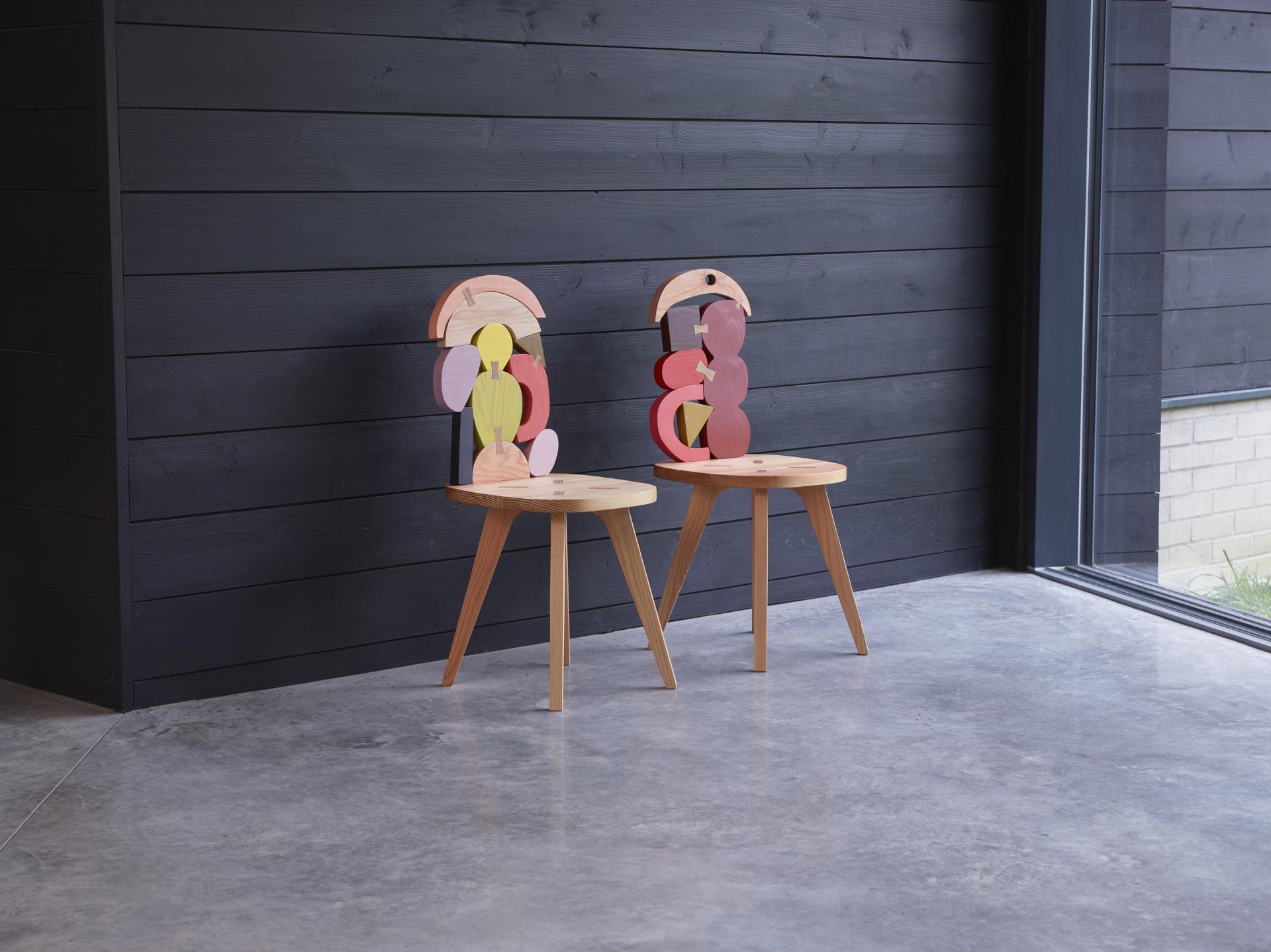 Two contemporary wooden chairs with playful abstract backrests, designed by SCP and Donna Wilson, against a black wall.