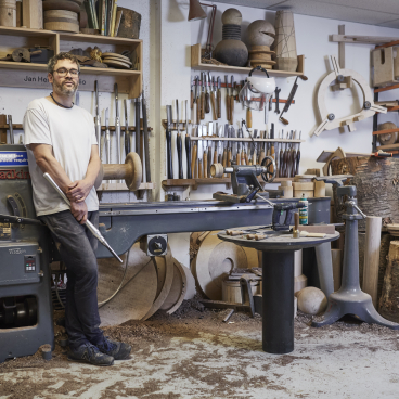 Craft artisan standing in a woodworking studio surrounded by tools, wood pieces, and a lathe, showcasing craft knowledge and creativity.
