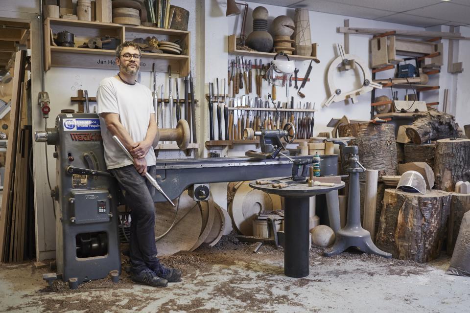 Craft artisan standing in a woodworking studio surrounded by tools, wood pieces, and a lathe, showcasing craft knowledge and creativity.