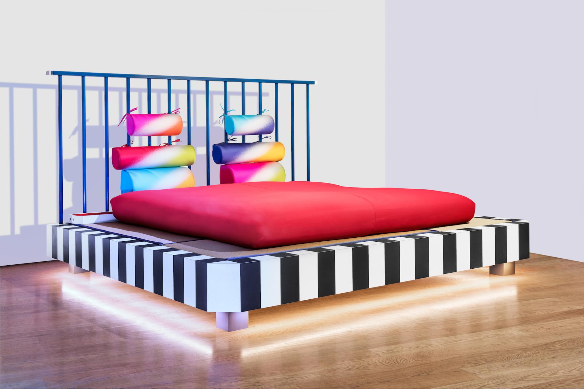Vibrant Memphis-inspired bed design featuring a red mattress and colorful cylindrical pillows, showcased at Fuorisalone Design City Edition 2021.