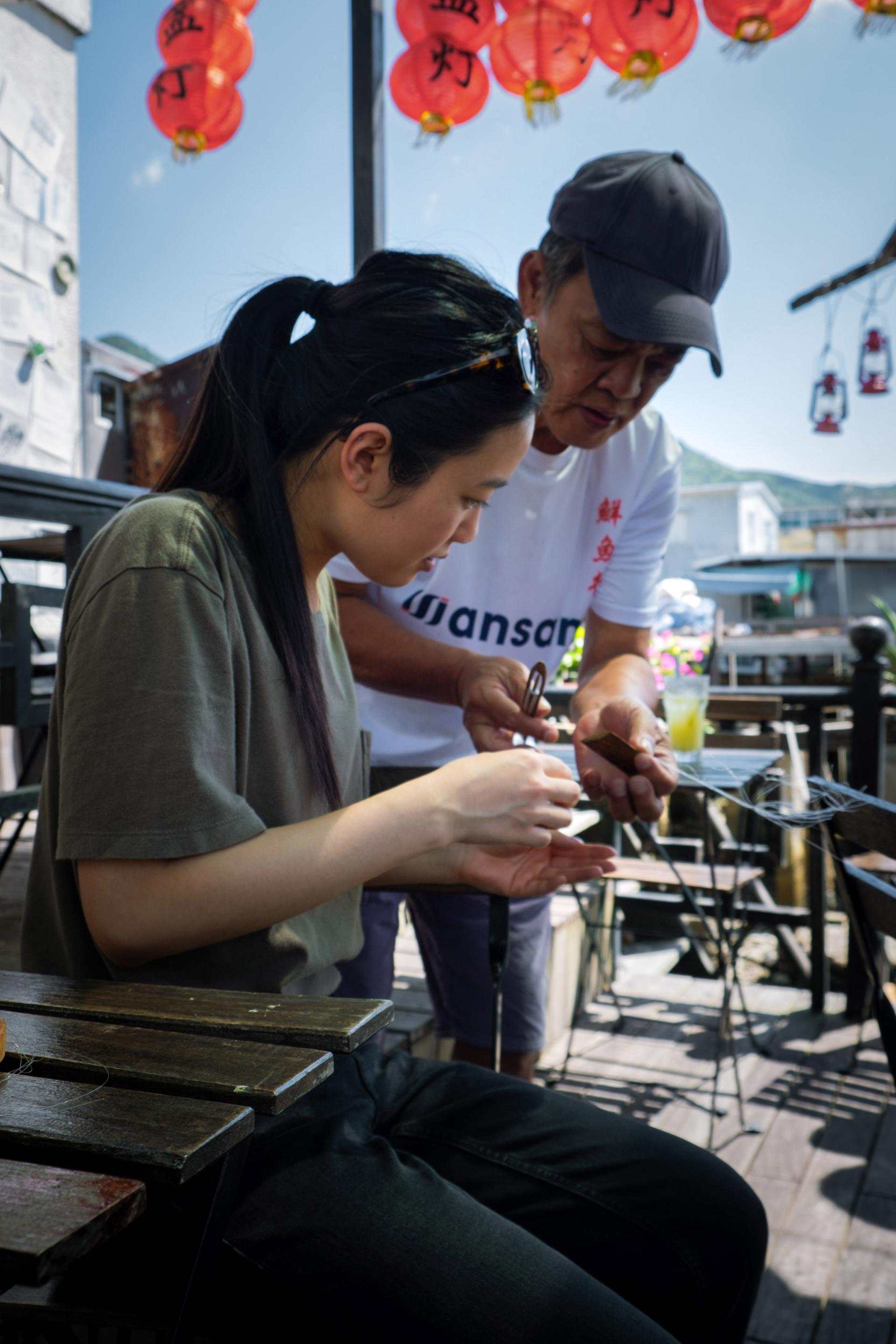 A woman learns local craft techniques from a mentor, surrounded by vibrant lanterns and a bustling market atmosphere.