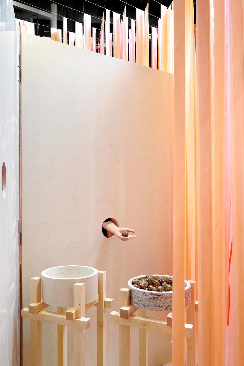 A hand holds a food item through a circular opening, surrounded by soft, colorful curtains and minimalist stone vessels.
