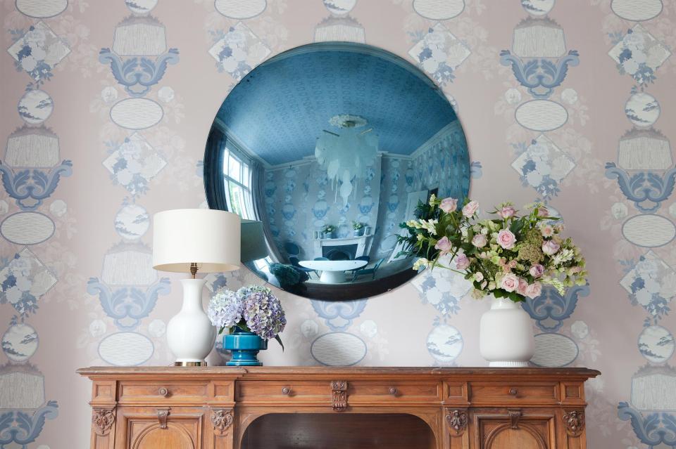 Elegant interior featuring a round mirror, vintage wooden furniture, and a floral arrangement against a patterned wallpaper backdrop.