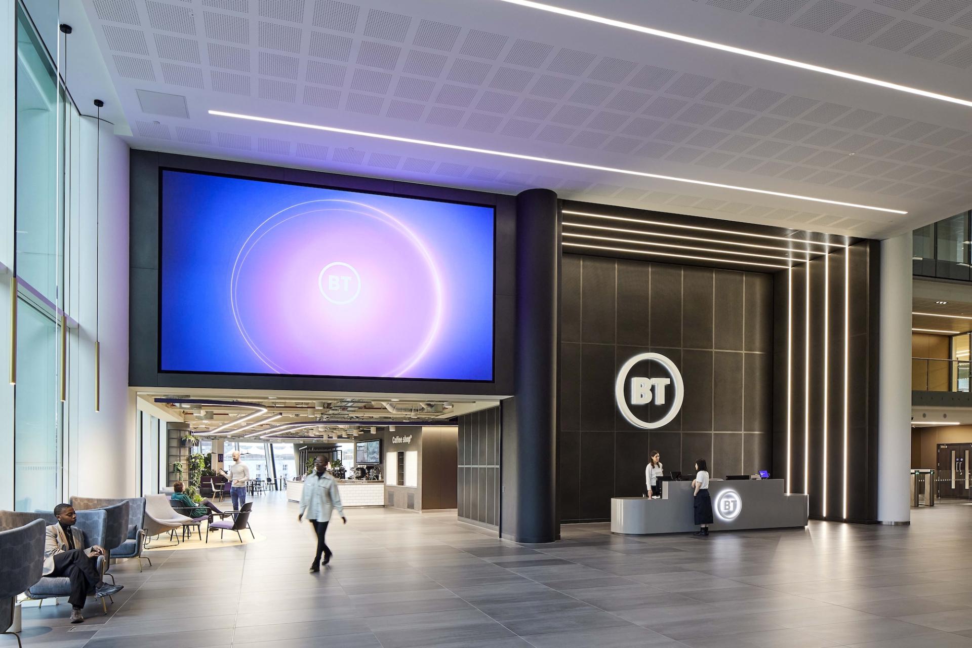 Modern office reception area featuring BT branding, digital display, and people engaged in conversation.