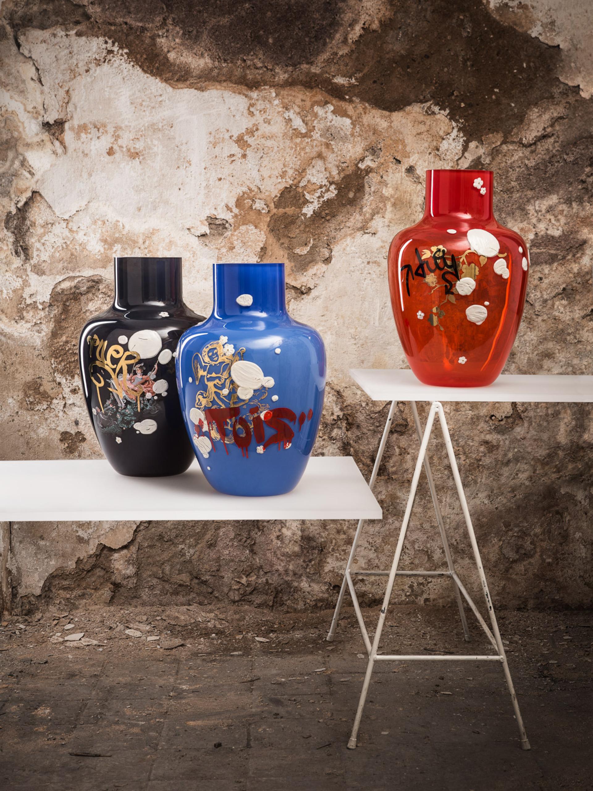 Artistic glass vases in blue, black, and red showcased on minimalist white tables against a textured wall backdrop.