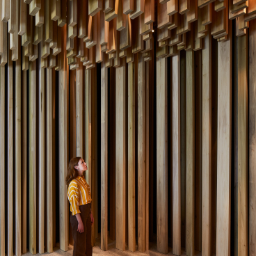 Visitor gazes upward at the intricate wooden structures of the American Tulipwood Pavilion designed by David Adjaye.