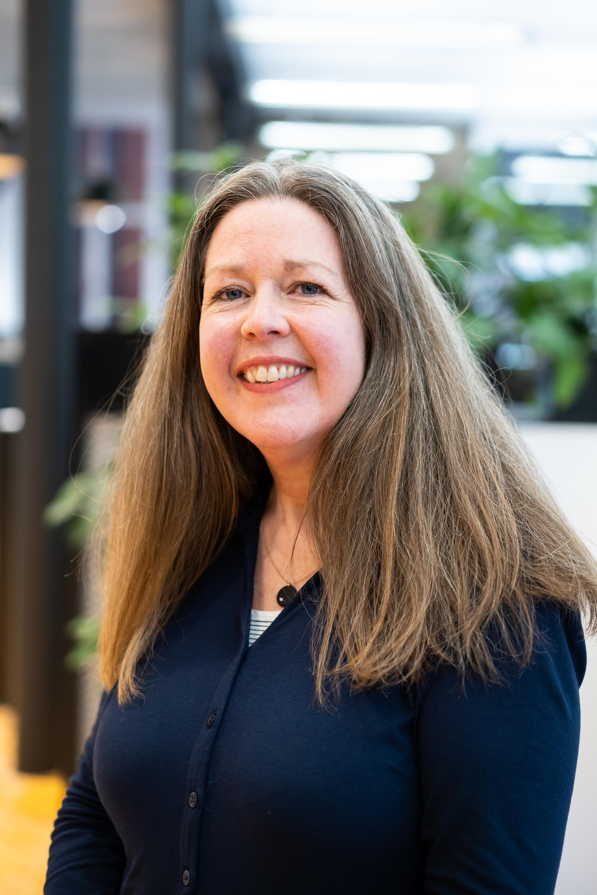 Professional woman with long hair smiles in a bright, green-plant-filled workspace, emphasizing sustainability in the built environment.
