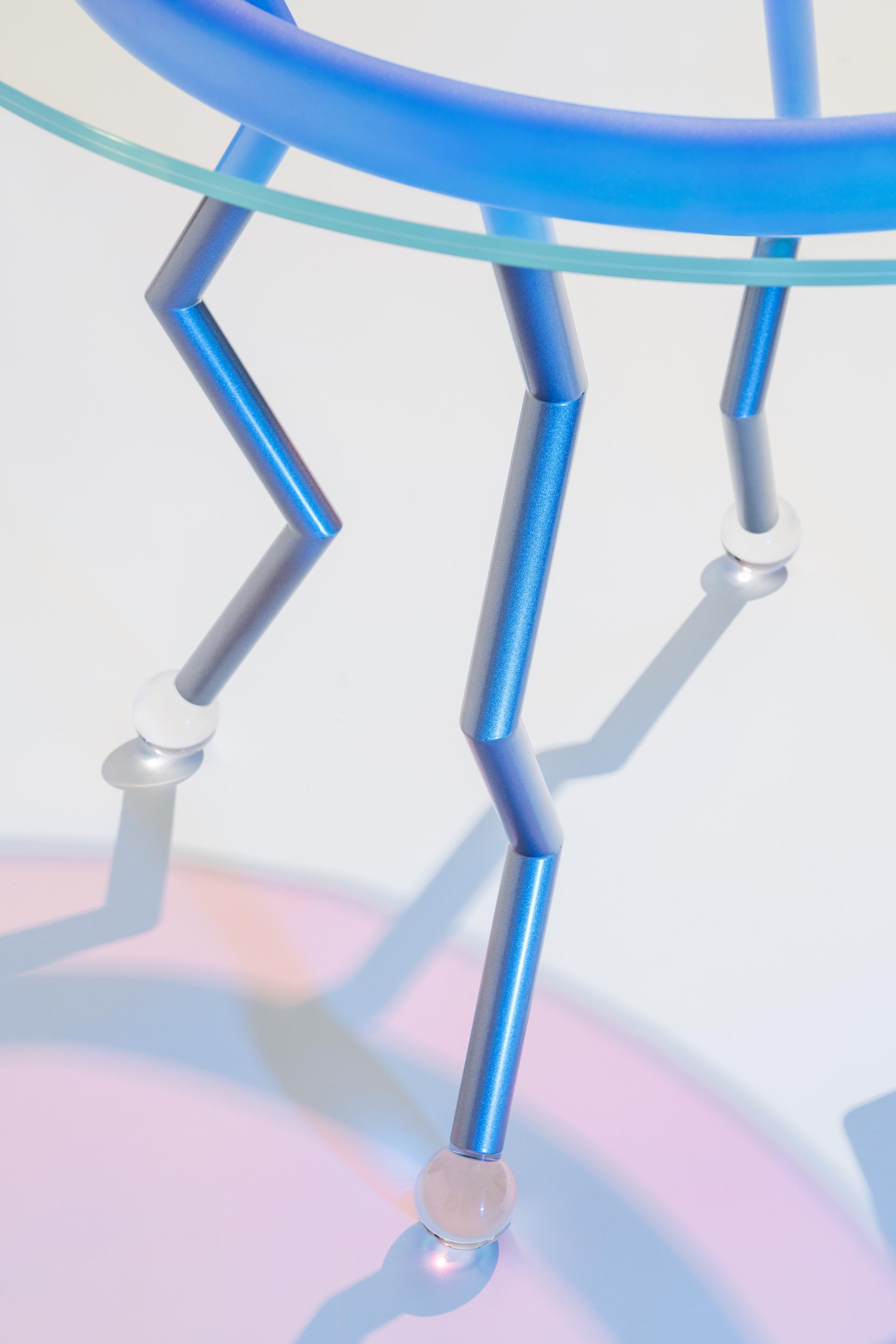 Colorful blue and silver table legs support a glass top, showcasing playful design elements from the Memphis movement.