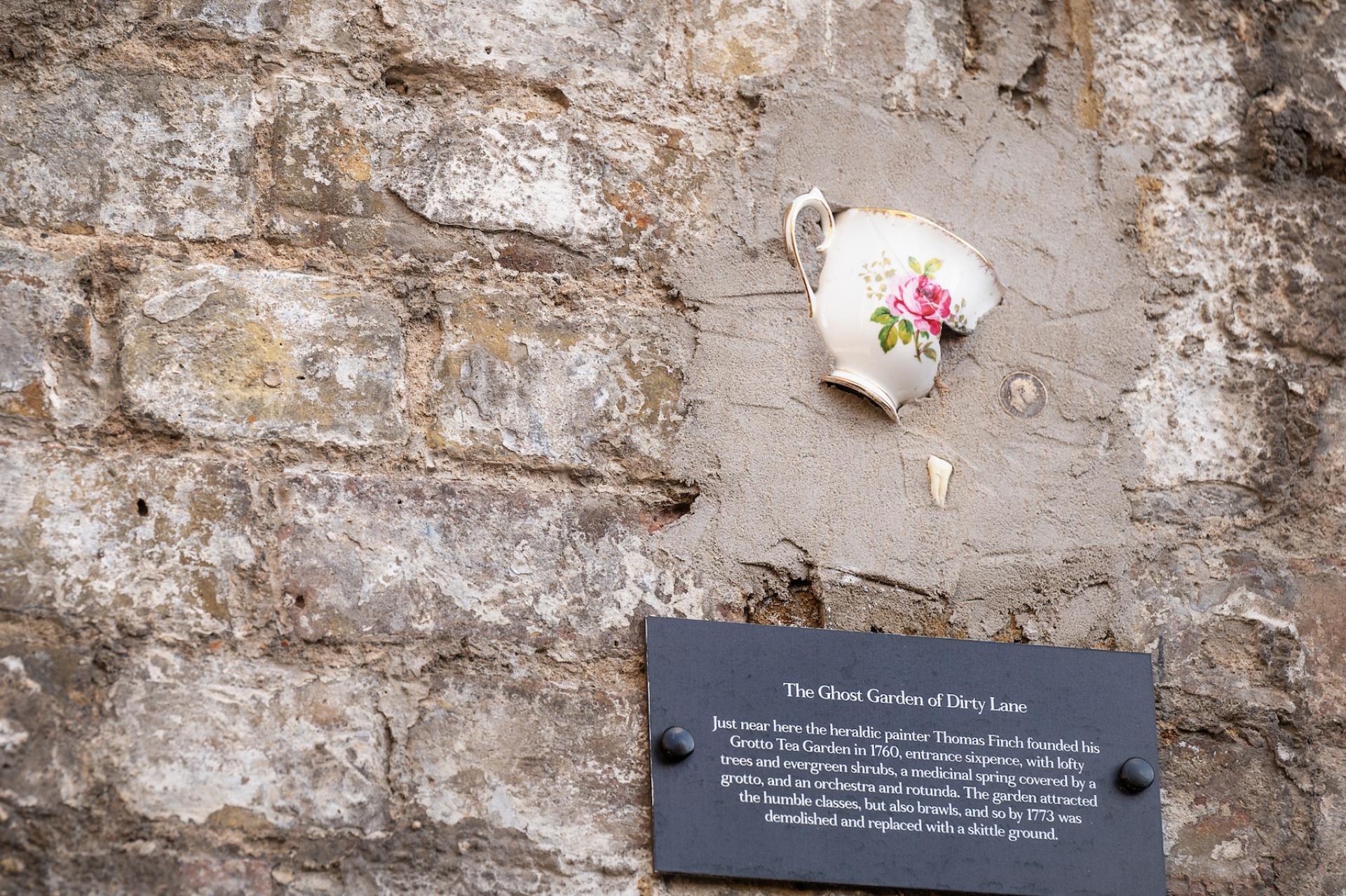 Broken teacup mounted on a weathered brick wall, accompanied by an informational plaque about the Ghost Garden of Dirty Lane.