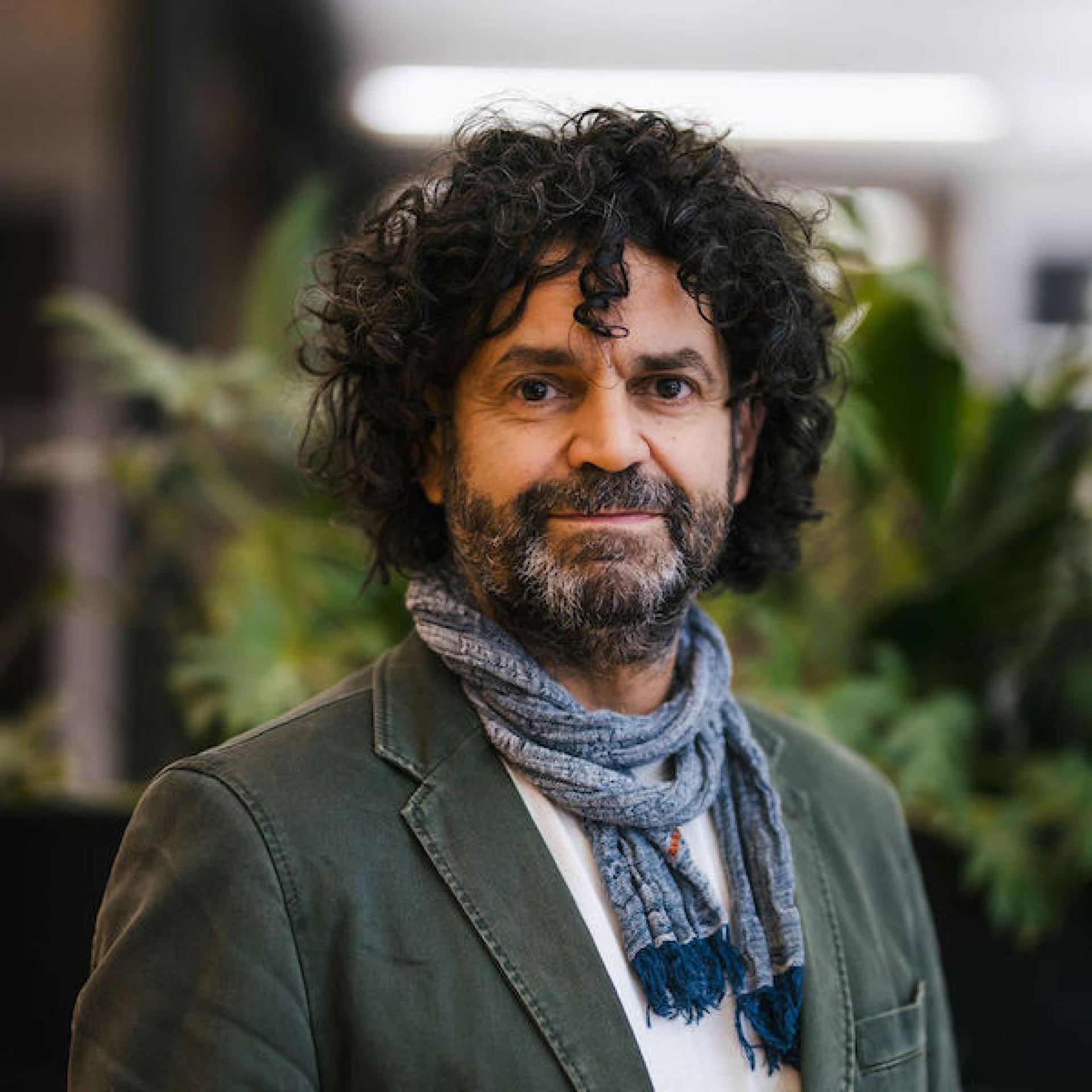 Man with curly hair and beard, wearing a scarf and jacket, stands in a green indoor setting related to Glasgow property discussions.