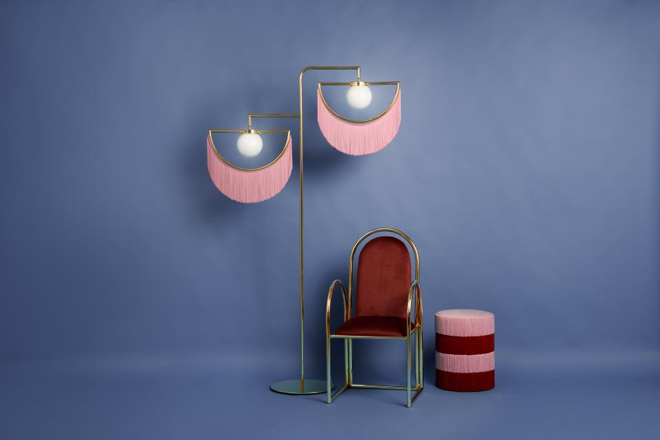A vintage-inspired seating area featuring a pink fringed lamp, red upholstered chair, and striped stool against a blue background.