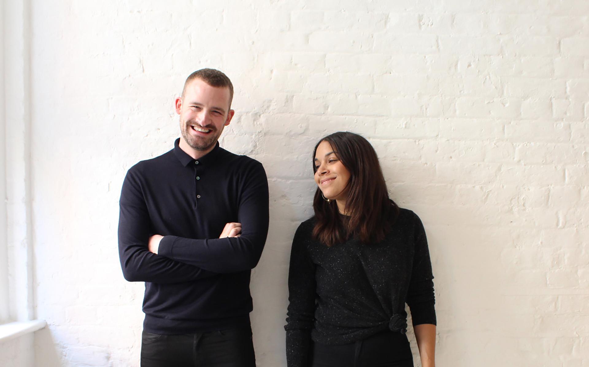 Cristina Rivadulla-Rey and Dean Thomas smiling together against a white brick wall, showcasing their creative consultancy work.