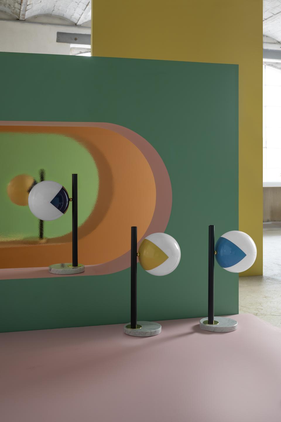 Colorful 'eyeball' lamps on sleek black stems showcase modern design against a vibrant geometric backdrop.
