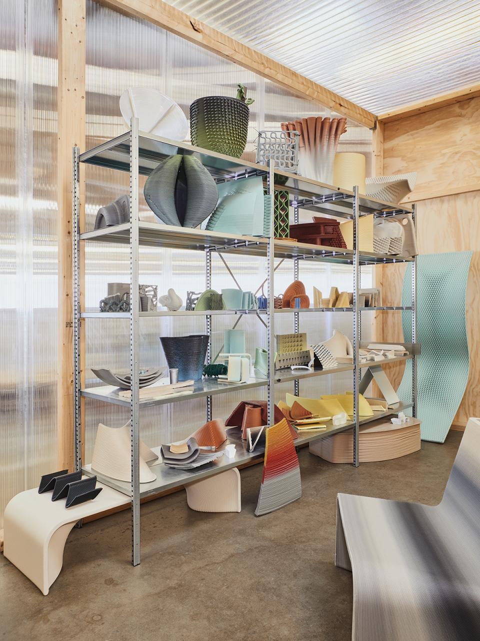 Colorful 3D-printed objects and the Ermis chair displayed on shelves in a minimalist design studio setting.