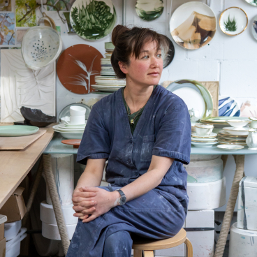 Reiko Kaneko, a ceramicist, seated in her studio surrounded by colorful hand-crafted plates and ceramic art.