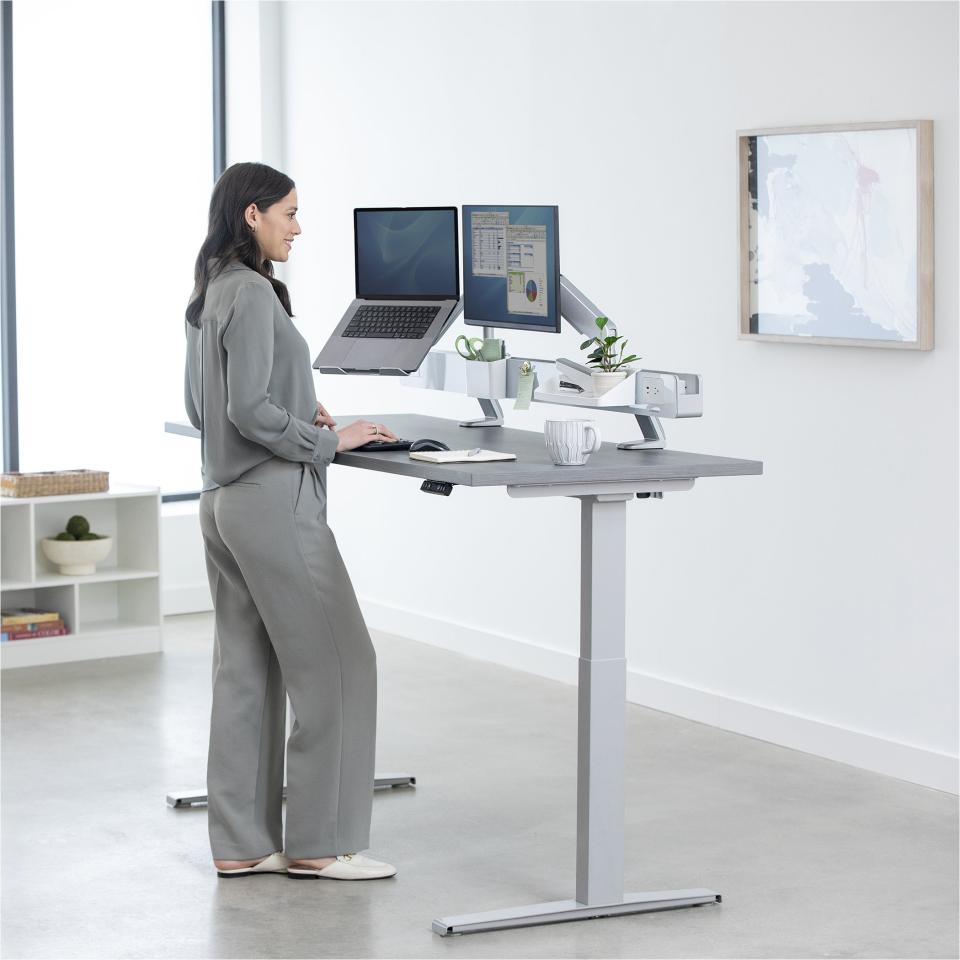 Standing woman working at an adjustable height desk with dual monitors, laptop, and workspace accessories for optimal productivity.